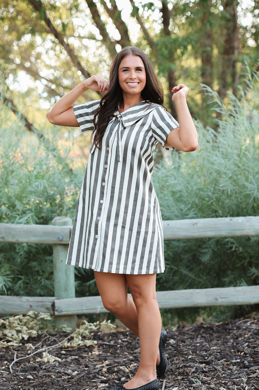 A woman wearing a short-sleeve button-front dress with vertical black and white stripes, posing outdoors in front of a wooden fence and green foliage.